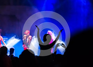 Concert crowd attending a concert, people silhouettes are visible, backlit by stage lights. Raised hands and smart phones are visi