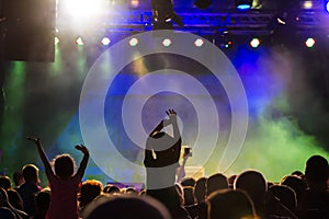 Concert crowd attending a concert, people silhouettes are visible, backlit by stage lights. Raised hands and smart phones are visi