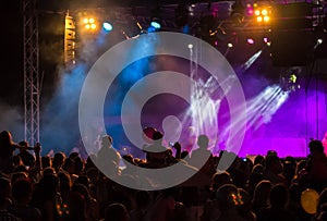 Concert crowd attending a concert, people silhouettes are visible, backlit by stage lights. Raised hands and smart phones are visi