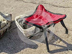 On the concrete floor there is a chair of three legs with a red seat for fishing. Red chair for fishing.
