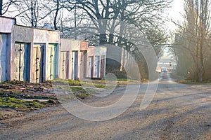 Concrete garages with colorful metal doors in morning sunlight along a road through alley of trees