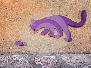 a concrete orange wall with purple wooden cat stalking a small mouse , funny purple cat chasing a scaredy small mouse in a cement