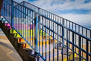 Concrete steps painted bright rainbow colours with a blue railing infront of a beach with sand. the sky is blue with white clouds