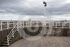 Concrete walkway with white metal painted railings and floodlights at the end of a pier with the sea in the background