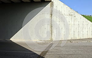 Concrete wall of an underpass divided in two by the shadow of the bridge with grass and sky on th right. Cement sidewalk and road
