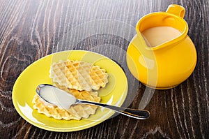 Condensed milk in spoon on cookie in yellow saucer, pitcher with condensed milk on wooden table