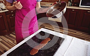 Confectioner chocolatier wearing pink apron pouring melted chocolate on marble surface on the background of wooden kitchen