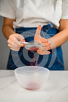 Confectioner sprinkling red powder over empty plastic cooking bowl on kitchen table