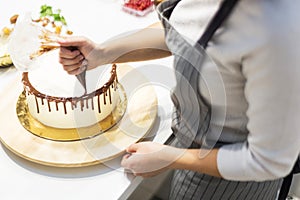 A confectioner squeezes liquid chocolate from a pastry bag onto a white cream biscuit cake on a wooden stand. The