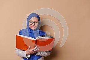 Confiden Muslim woman, wearing strict outfit and a blue hijab, reads a hardcover book on isolated beige background