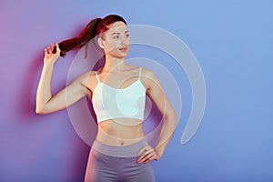 Confident active healthy woman in sports clothing with one hand on hip posing isolated over clor background, tocghing her ponytail