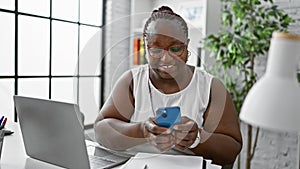 Confident african american business woman, working with laptop and smartphone, smiling boldly at the office interior