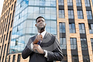 Confident african american businessman adjusting necktie, standing against office center in urban area, free space
