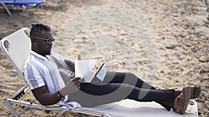 Confident African American freelancer lying on sunbed persuading client in video chat on tablet analyzing paperwork
