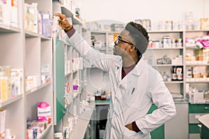 Confident African American man pharmacist standing in interior of pharmacy and searching the drug on shelves in pharmacy