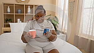 Confident african american woman comfortable in her skin, relaxing in bed, drinking her morning coffee, texting on her phone, her