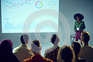 Confident African-American woman, expert with stands in front of audience, giving lecture with data chart backdrop in