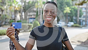 Confident african american woman, radiating happiness, flaunting her credit card on a busy city street, embodying financial