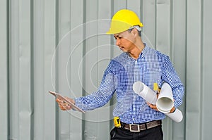 Confident architect. Handsome young man in hardhat holding blueprint and using tablet while standing outdoors