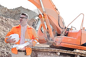 Confident architect holding hardhat and blueprints at construction site