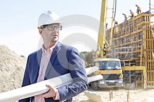 Confident architect holding rolled up blueprints at construction site