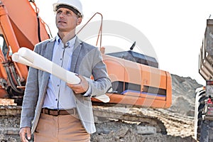 Confident architect looking away while holding blueprints at construction site