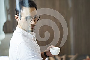 Confident Asian businessman standing behind glass drinking coffee looking down at the city