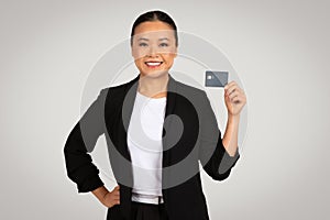 A confident Asian businesswoman in a black suit holds up a credit card with a poised smile