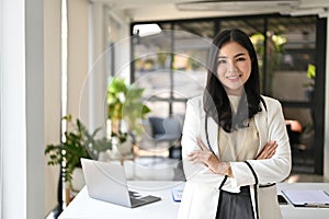 Confident Asian businesswoman in white suit stands with arms crossed in the office