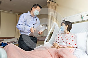 Confident Asian doctor with face mask explaning to cheerful female patient on medical condition lying on bed in hospital