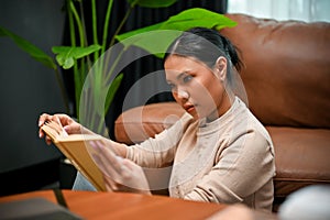 Confident Asian female focused reading a book in her modern vintage living room