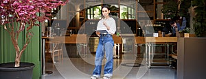 Confident asian woman, cafe manager or employee, standing near entrance with tablet, looking thoughtful