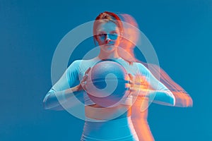 Confident athletic girl basketball player holding ball looking at camera on blue background. Long exposure, motion blur