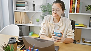 Confident and attractive young hispanic woman enjoying coffee while balancing business on her laptop at the office