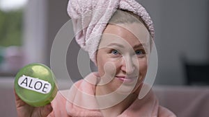 Confident attractive young woman in hair towel and bathrobe applying moisturizer on face looking at camera showing Aloe