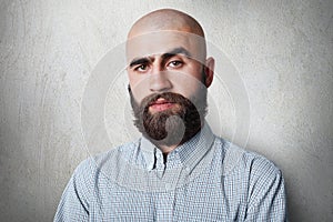 A confident bald male with thick black eyebrows and beard wearing checked shirt having gloomy expression posing against white back