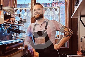 Barista with stylish beard and hairstyle wearing apron smiling and looking sideways while leaning on a counter in the photo