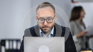 Confident bearded man in eyeglasses working at modern office on laptop sitting at desk workplace