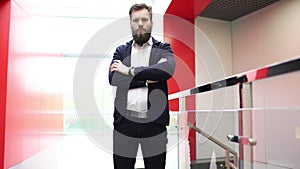 Confident bearded man is standing in front of the camera near glass fence in modern building crossing his hands. Close