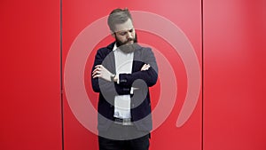 Confident bearded man is standing in front of the camera near red wall, crossing his hands and looking at watch. Close