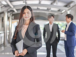 Confident beautiful Asian business woman in formal suit holding computer laptop, standing in front of her team, smiling and