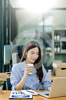 Confident beautiful Asian businesswoman typing laptop computer and digital tablet while holding coffee