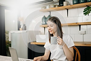 Confident beautiful Asian businesswoman typing laptop computer and digital tablet while holding coffee at home