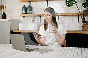 Confident beautiful Asian businesswoman typing laptop computer and digital tablet while holding coffee at home