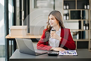 Confident beautiful Asian businesswoman typing laptop computer and digital tablet while holding coffee at modern office