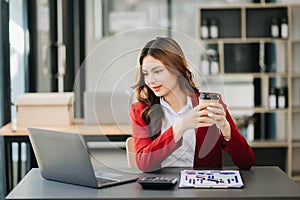 Confident beautiful Asian businesswoman typing laptop computer and digital tablet while holding coffee at modern office