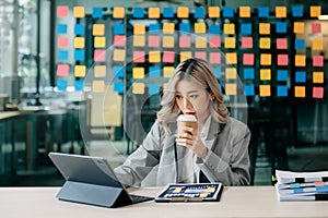 Confident beautiful Asian businesswoman typing laptop computer and digital tablet while holding coffee at office