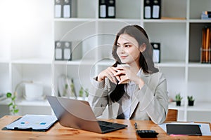 Confident beautiful Asian businesswoman typing laptop computer and digital tablet while holding coffee at office