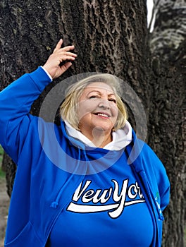 Confident beautiful mature woman stands with raised hands near a tree and smiles.