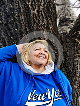 Confident beautiful mature woman stands with raised hands near a tree and smiles
